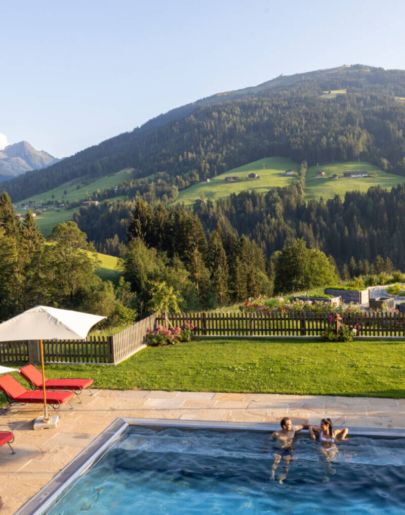 Ein Mann und eine Frau sitzen im Pool und reden miteinander - im Hintergrund sieht man den Garten und gegenüberliegenden Berg