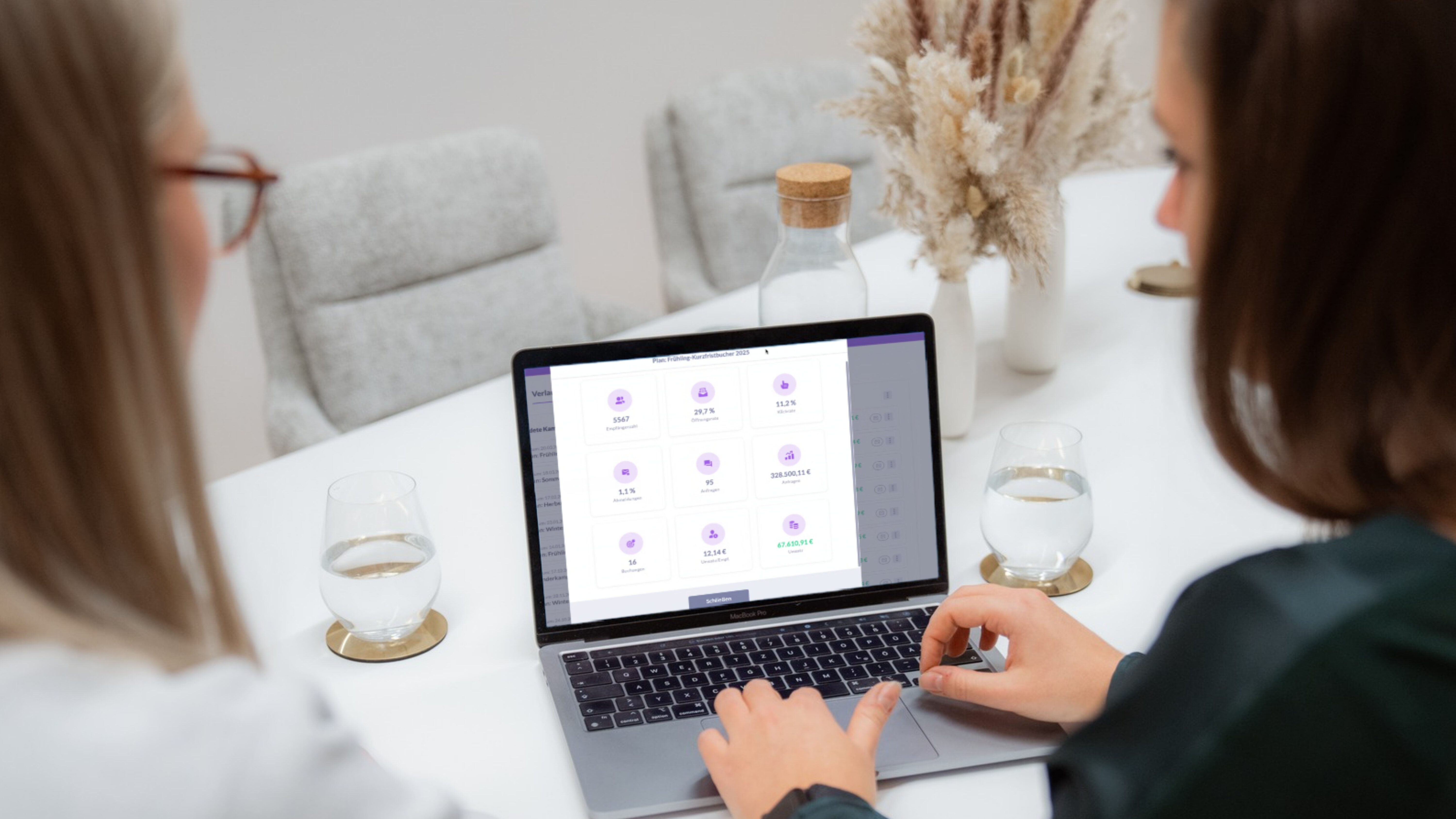 Zwei Frauen sitzen an einem hellen Tisch und blicken gemeinsam auf ein aufgeklapptes Laptop, dessen Bildschirm eine Software-Oberfläche mit mehreren runden Icons zeigt. Auf dem Tisch stehen zwei Gläser Wasser auf goldfarbenen Untersetzern sowie eine Karaffe; im Hintergrund ein Strauß getrockneter Gräser. 